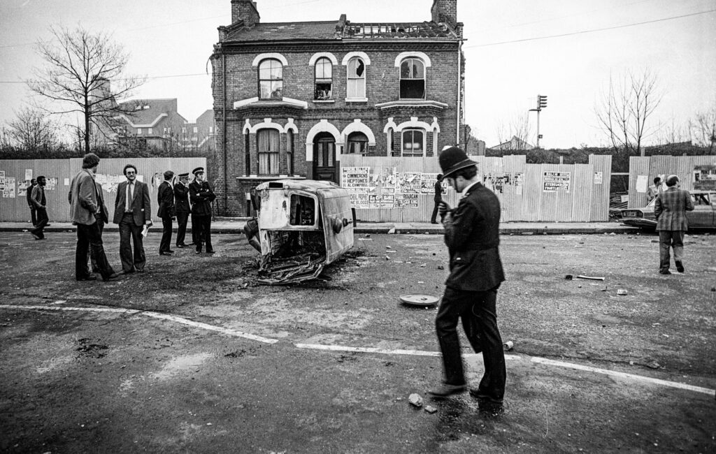 A burnt out police van lies on its side in Railton Road  while residents watch on from a nearby house