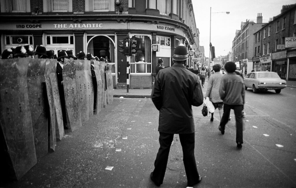 Police cordon off Railton Road by The Atlantic which was later destroyed buy rioters. Members of the public continue to shop in Coldharbour lane, despite the destruction only streets away.