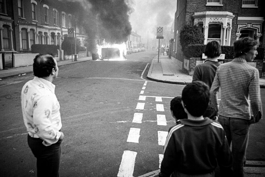Residents from nearby houses look on as a car burns.