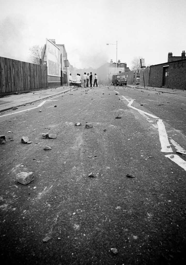 Rioters gather toward the end of a deserted and rock-strewn Railton Rd. To the right, an abandonded police van.