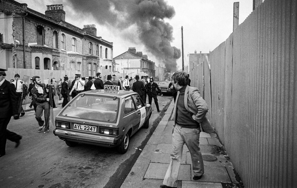Police and onlookers retreat rapidly as a vehicle is hit with a molotov cocktail and explodes